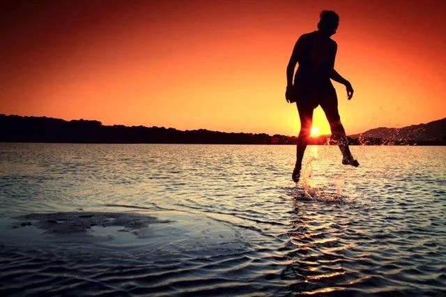 fotografia minimalista de pessoa pulando em água rasa em contraluz no pôr-do-sol